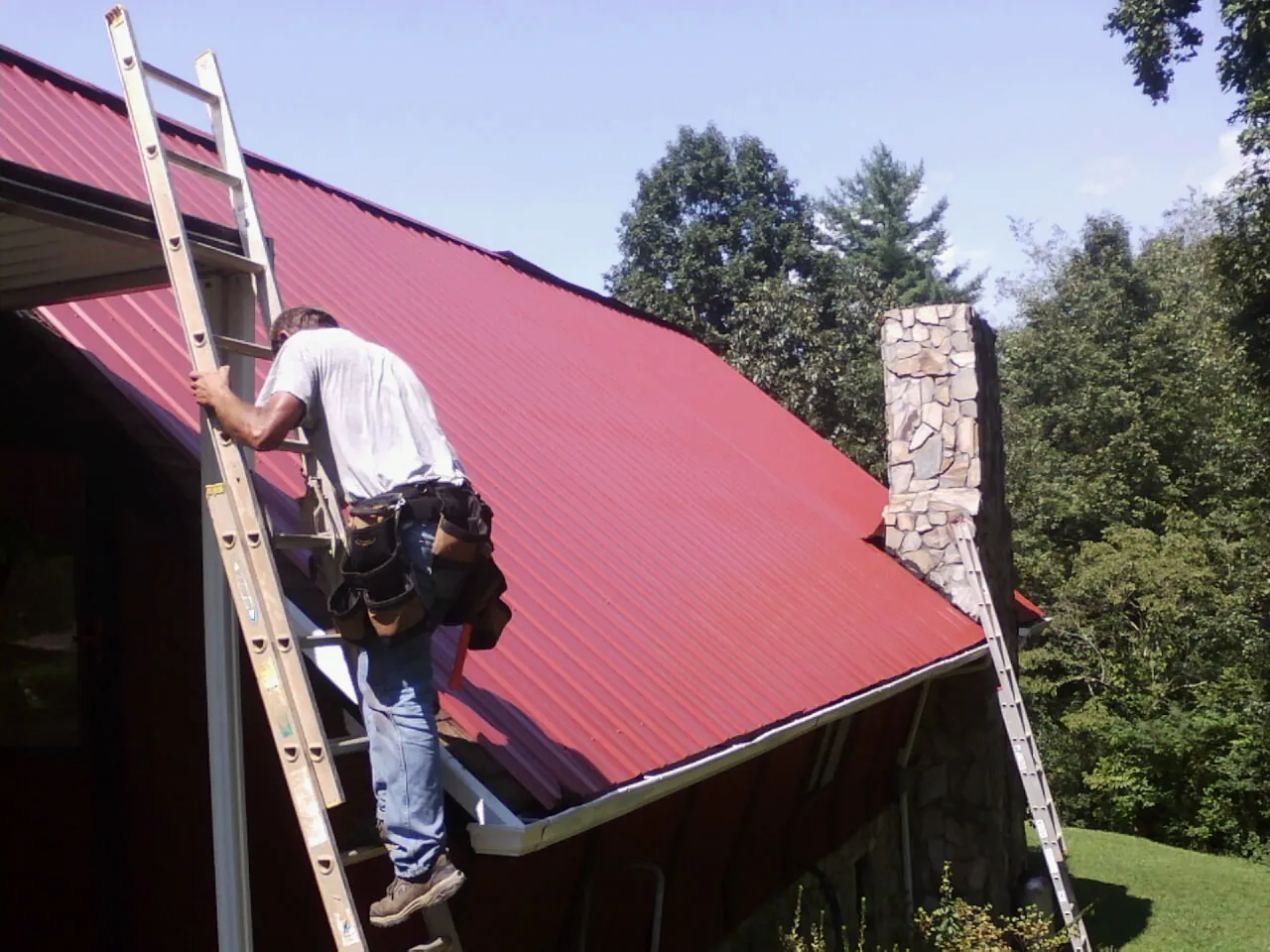 Roofer on a ladder beside a red metal roof and stone chimney — Soffit & Fascia Repair by Roofing Brothers in Thomasboro